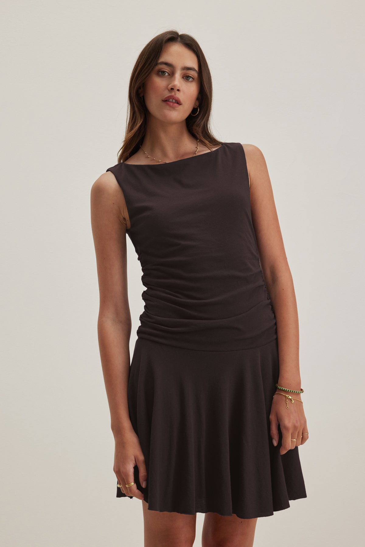 A woman stands wearing the sleeveless ALICIA DRESS by Velvet by Graham & Spencer, featuring a dark hue with a ruched bodice and flared skirt, exuding timeless elegance against a plain background.-38918664454337
