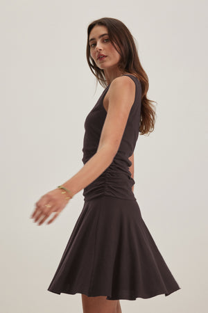 A woman with long brown hair in the ALICIA DRESS by Velvet by Graham & Spencer stands against a plain backdrop, exuding timeless elegance. Her slightly extended right arm captures classic sophistication that never fades.