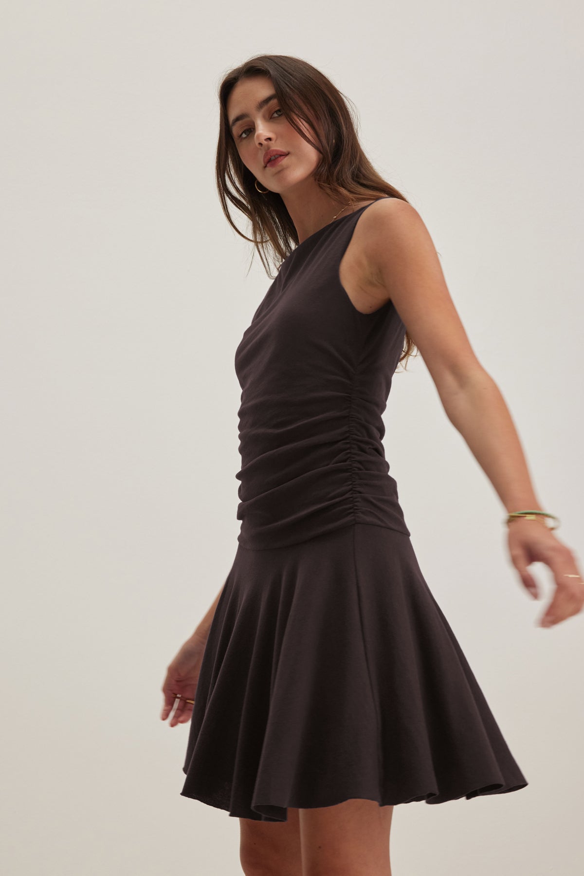 A woman in the ALICIA DRESS by Velvet by Graham & Spencer, a classic black sleeveless design with a ruched bodice and flared skirt, turning slightly against a plain background, embodies timeless style.-38918664585409