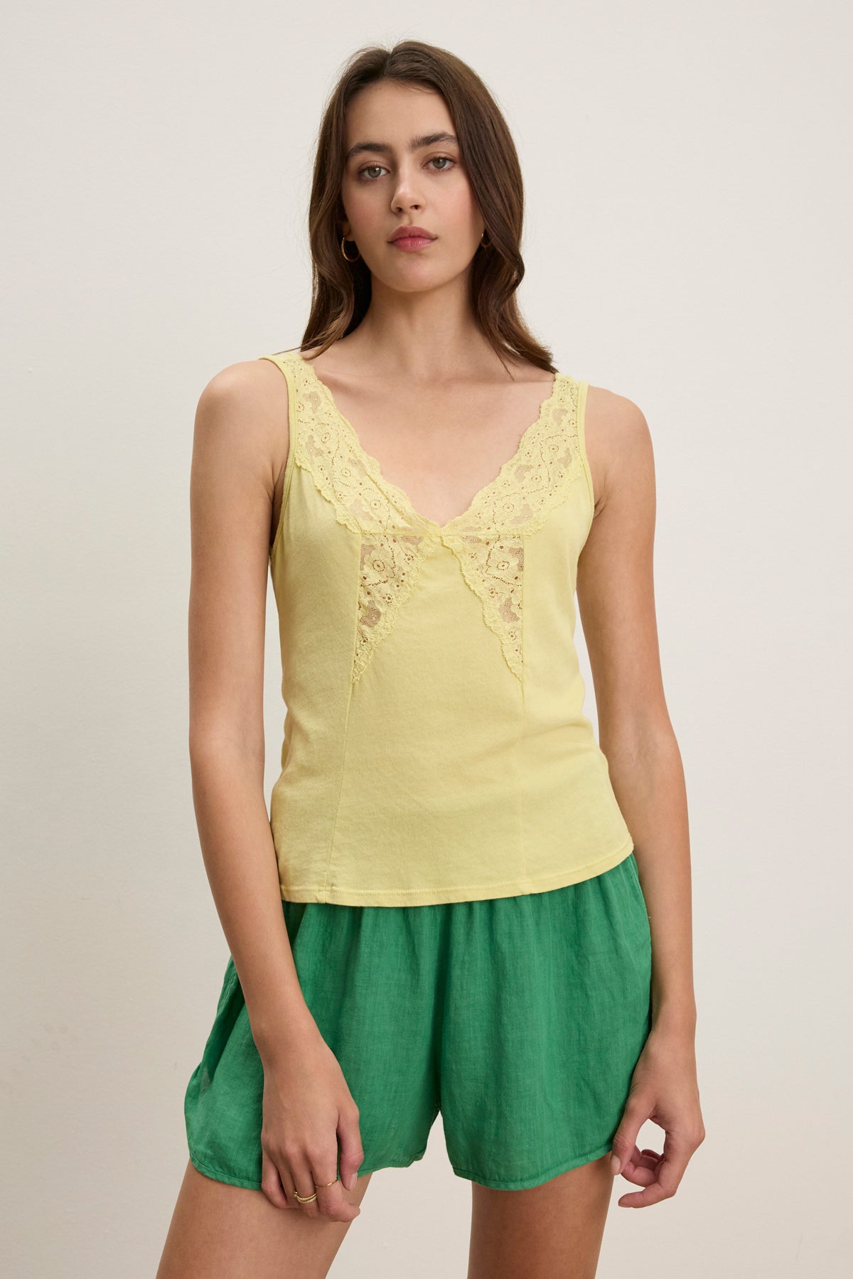 A woman stands against a plain background wearing the HANNAH LACE TANK TOP by Velvet by Graham & Spencer, a yellow V-neck sleeveless top with lace detailing, paired with green shorts.-39119886352577