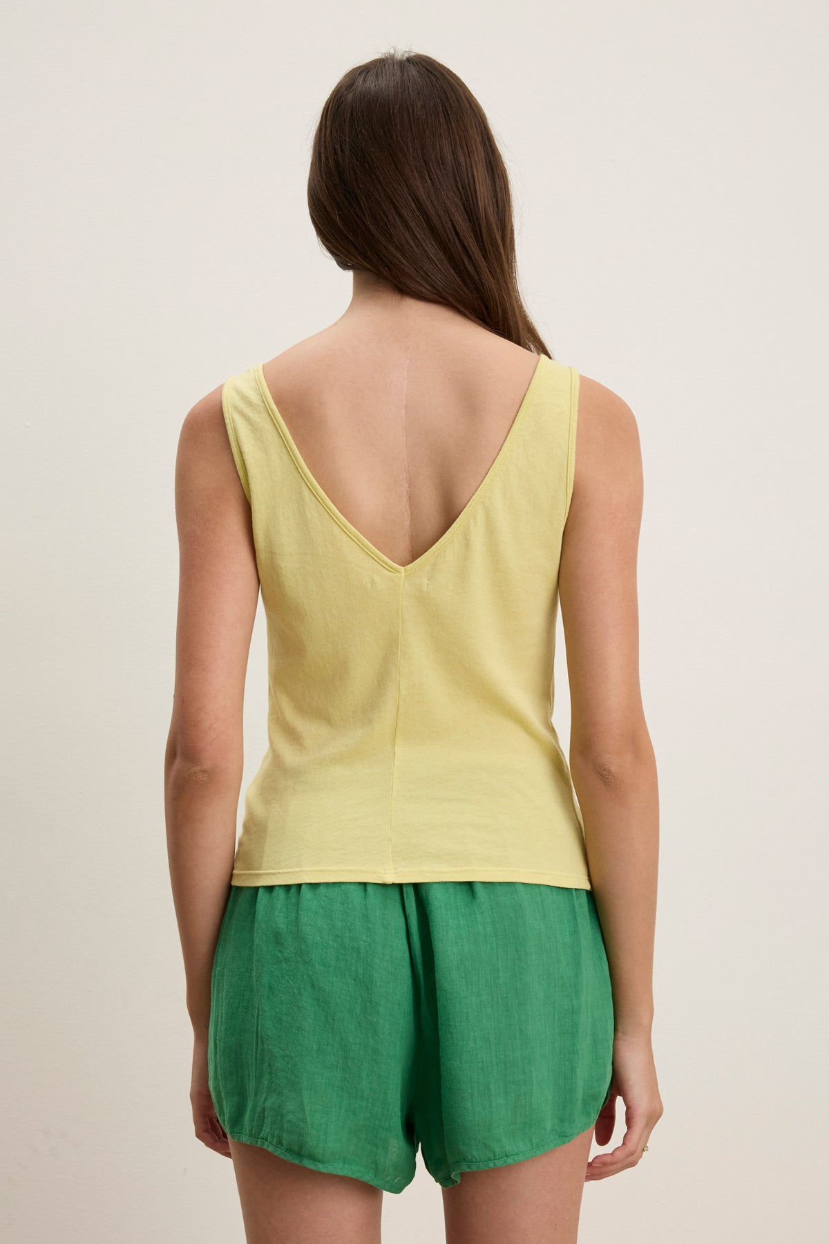 A woman with long brown hair, facing away from the camera, wears the HANNAH LACE TANK TOP by Velvet by Graham & Spencer—a sleeveless yellow V-back top with a V-neckline—paired with green shorts against a plain light background.-39119886090433