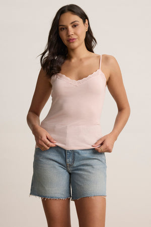 A woman stands facing the camera against a plain background, wearing the Velvet by Graham & Spencer INDIE LACE CAMI in pink paired with light blue denim shorts.