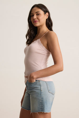 A woman stands sideways against a plain background, wearing the Velvet by Graham & Spencer INDIE LACE CAMI in light pink with lace details and denim shorts, one hand in her pocket.