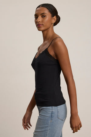Woman in the INDIE LACE CAMI by Velvet by Graham & Spencer—a black lace-detailed tank top—paired with light blue jeans, standing sideways against a plain background.