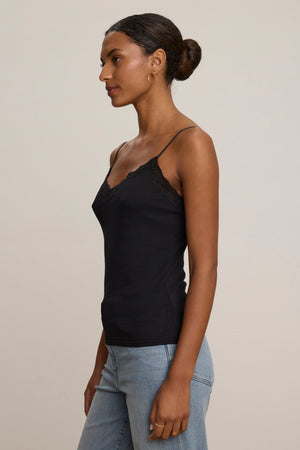A woman stands sideways against a plain beige background wearing the INDIE LACE CAMI by Velvet by Graham & Spencer with black lace detailing, paired with light blue jeans.
