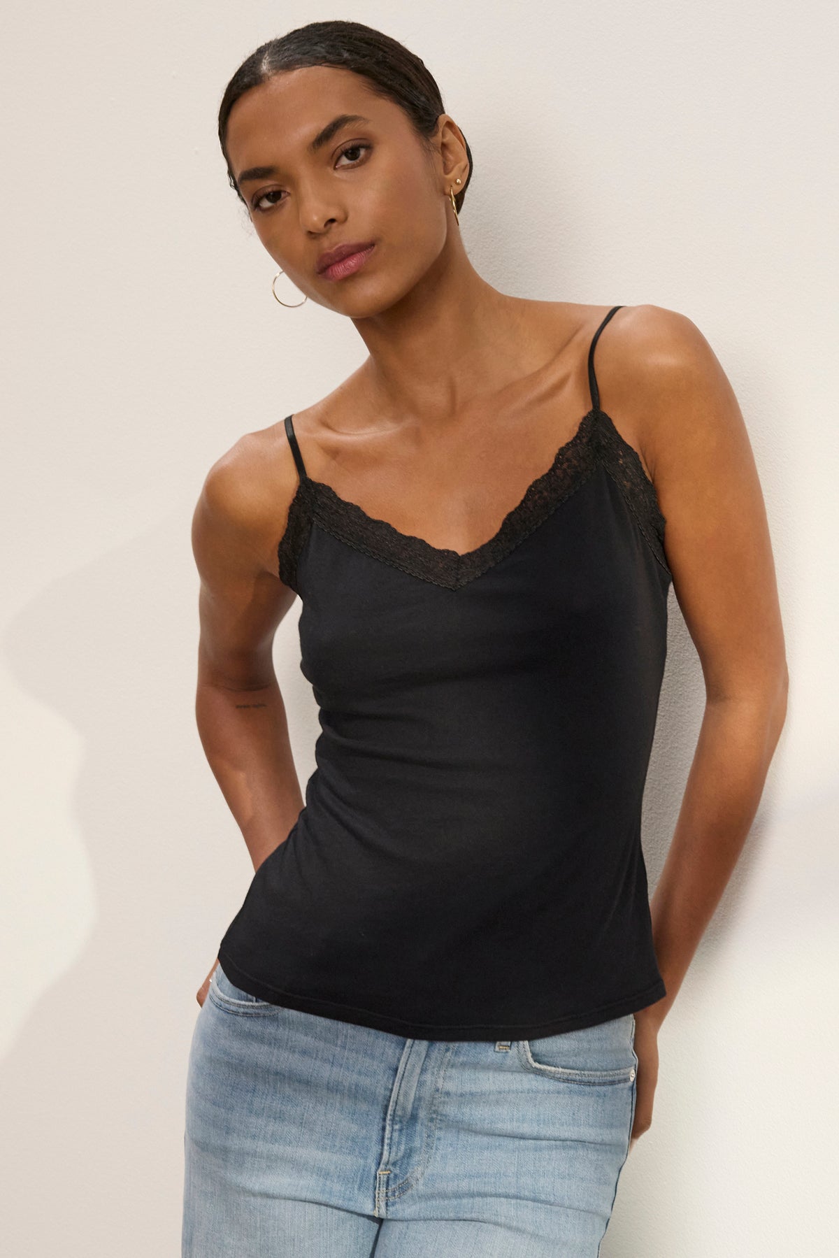   A woman wearing the INDIE LACE CAMI by Velvet by Graham & Spencer, a black tank top with delicate lace details, pairs it with light blue jeans and stands against a plain, light-colored wall. 