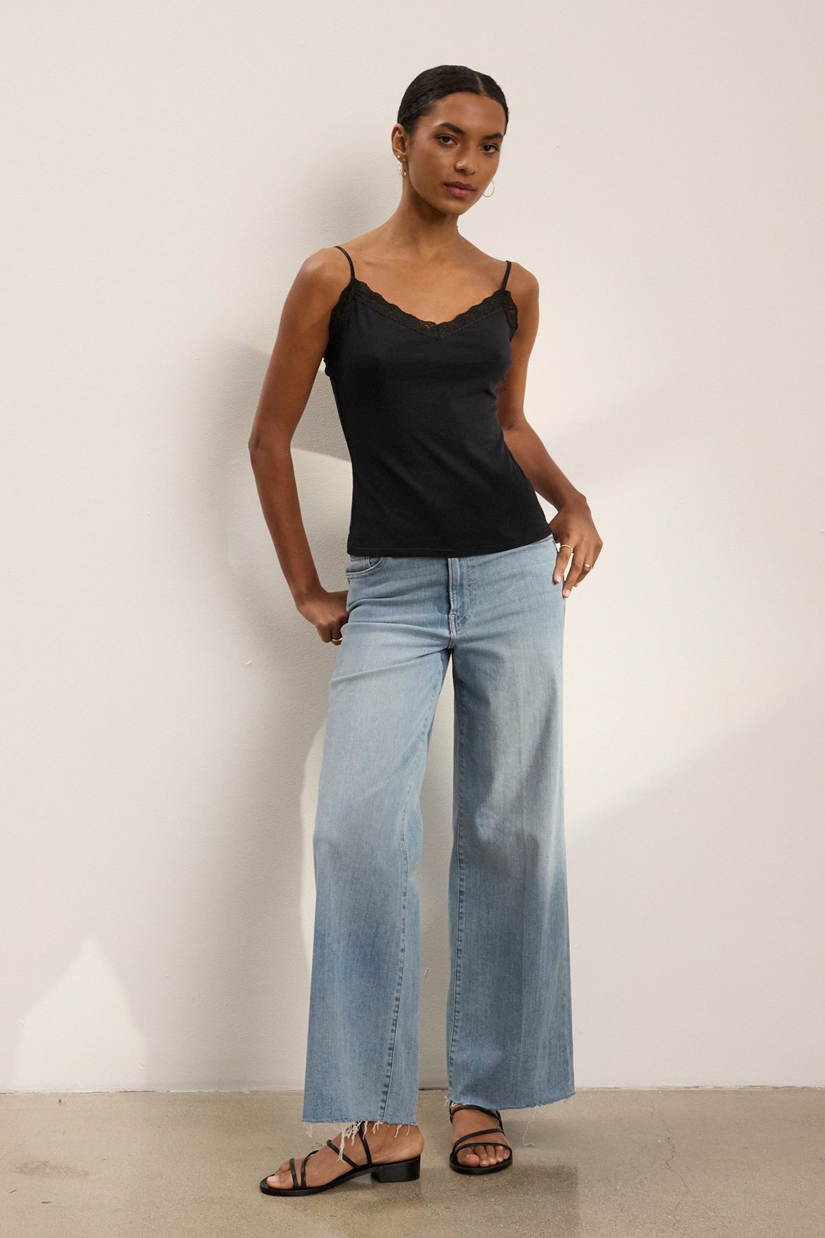 A woman stands against a plain wall wearing the Velvet by Graham & Spencer INDIE LACE CAMI, light blue wide-leg jeans, and black sandals.-39261838180545