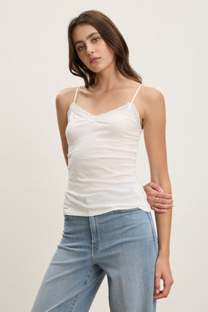 A woman models the INDIE LACE CAMI by Velvet by Graham & Spencer, featuring white lace detailing, paired with light blue jeans against a plain light background.