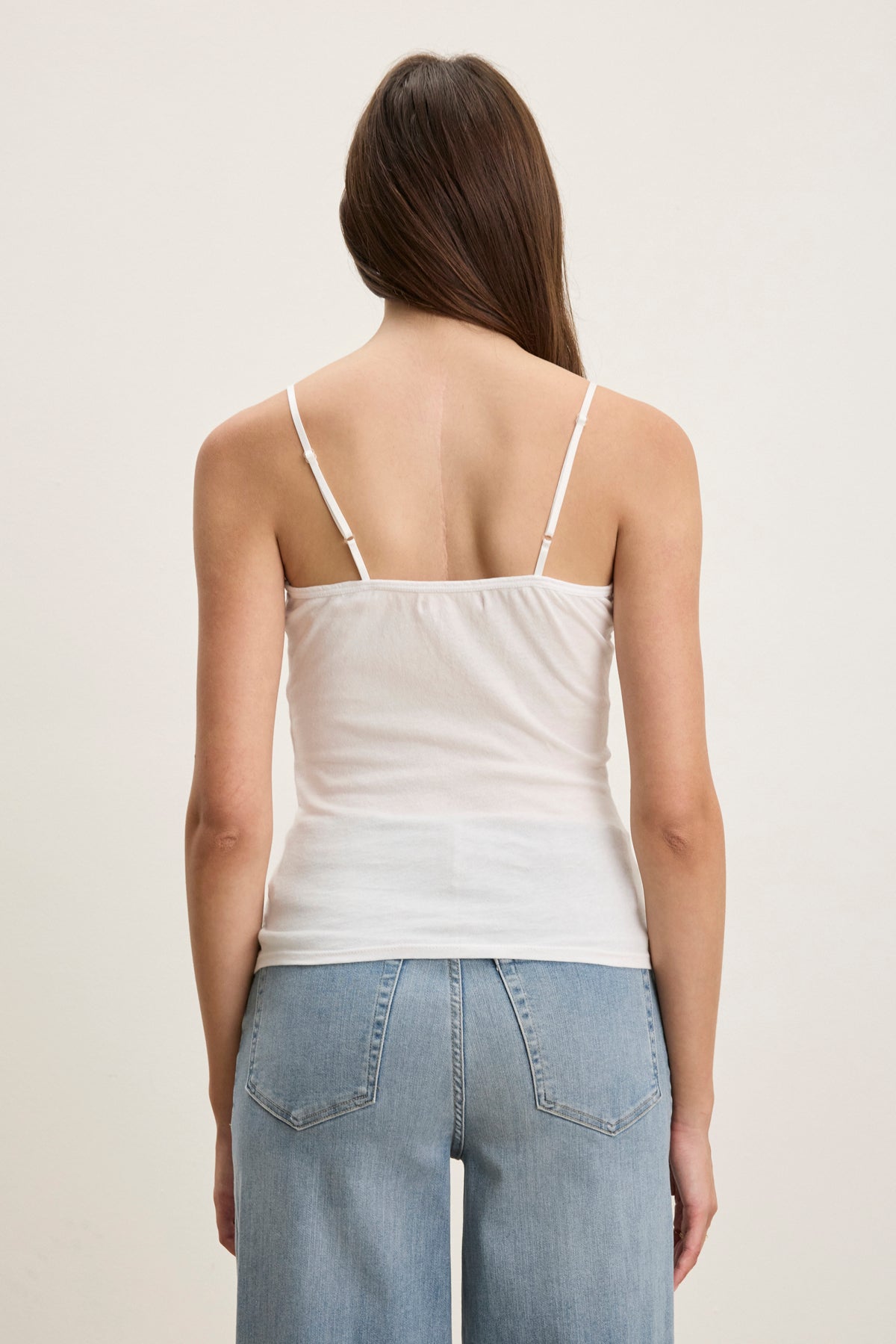 A person with long brown hair is seen from the back wearing the Velvet by Graham & Spencer INDIE LACE CAMI, a white tank top with lace detailing, paired with light blue jeans against a plain light background.-39079954874561