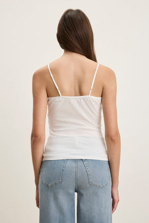 A person with long brown hair is seen from the back wearing the Velvet by Graham & Spencer INDIE LACE CAMI, a white tank top with lace detailing, paired with light blue jeans against a plain light background.
