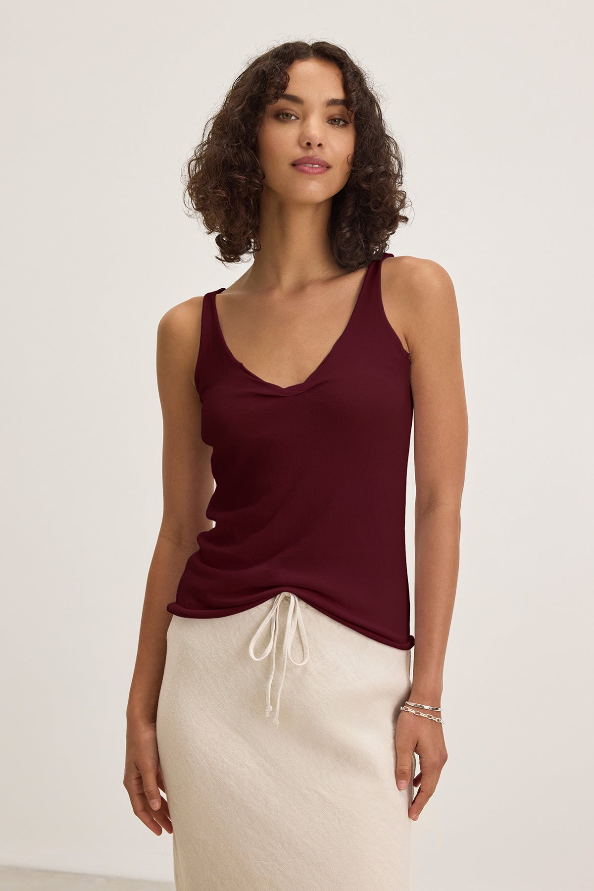 A woman stands against a plain background, wearing the KIM TANK TOP by Velvet by Graham & Spencer in burgundy and a light beige drawstring-waist skirt—effortless wardrobe essentials.-40009806577857