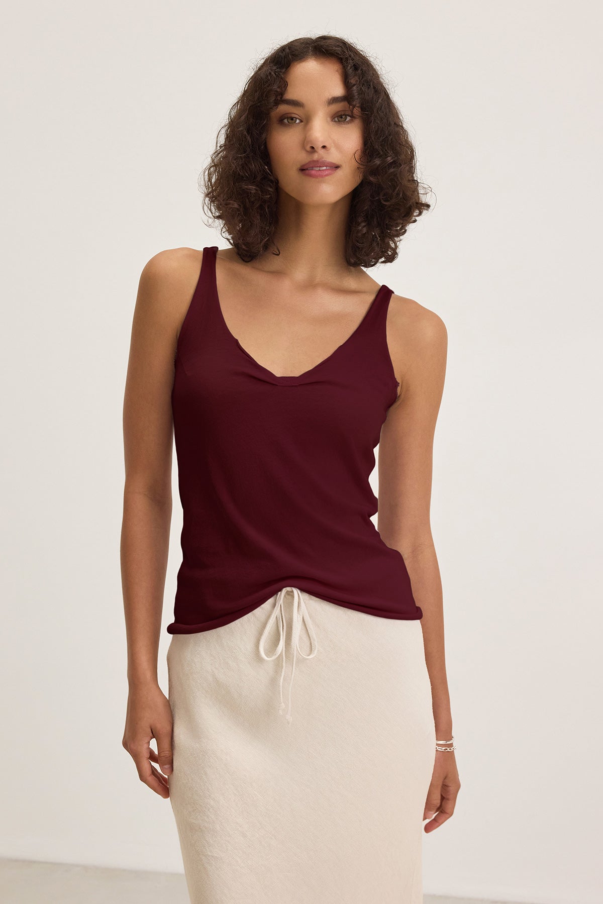 A woman wears the KIM TANK TOP by Velvet by Graham & Spencer—a sleeveless maroon V-neck top—paired with a cream skirt, standing against a plain light background.-40009806479553