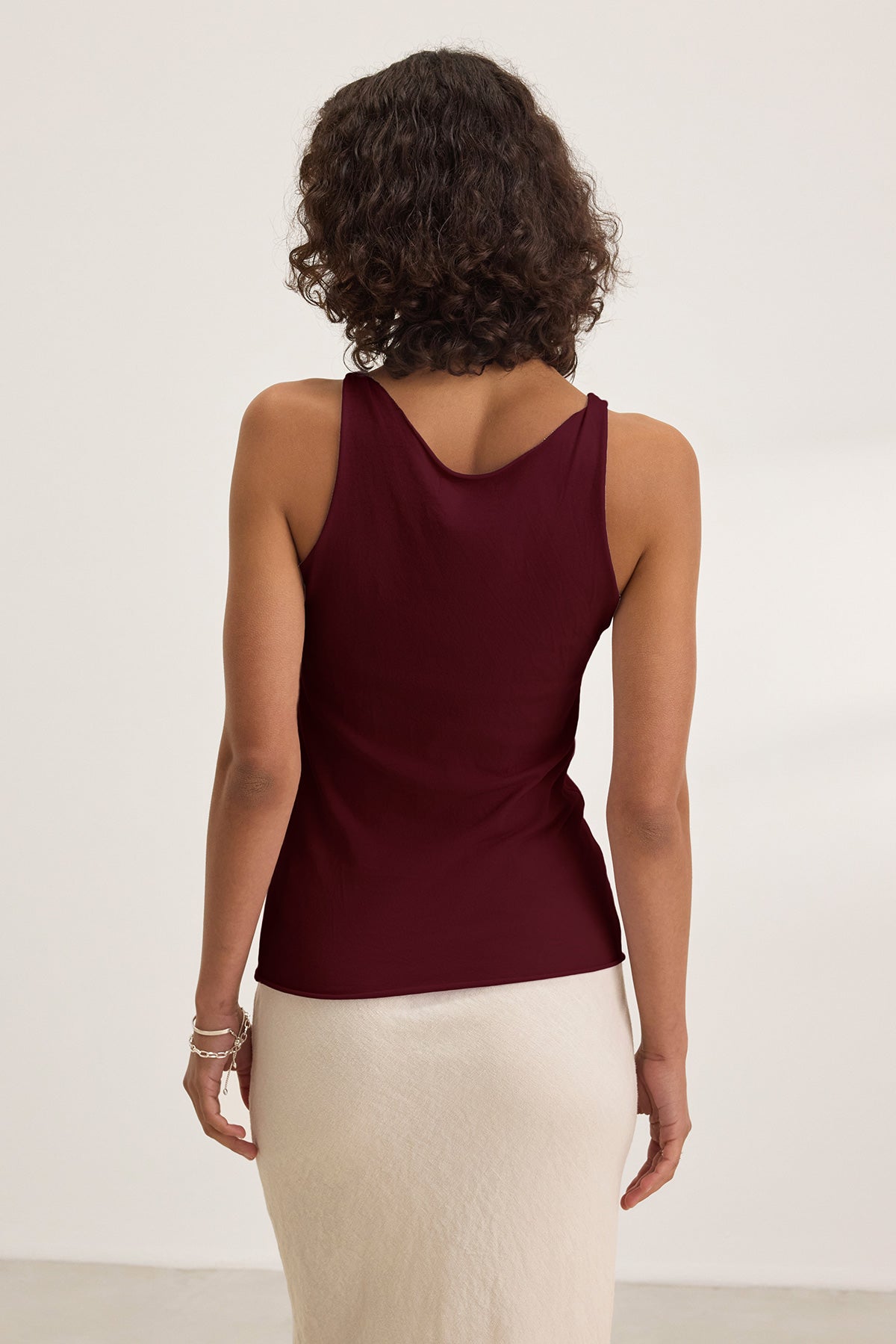 A woman with curly hair is seen from the back, wearing Velvet by Graham & Spencer's KIM TANK TOP—a sleeveless burgundy scoop-neck top—paired with a light skirt, against a plain light background.-40009806512321