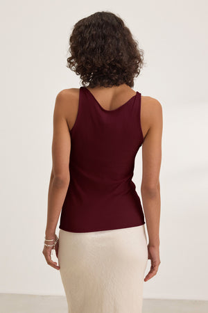 A woman with curly hair is seen from the back, wearing Velvet by Graham & Spencer's KIM TANK TOP—a sleeveless burgundy scoop-neck top—paired with a light skirt, against a plain light background.