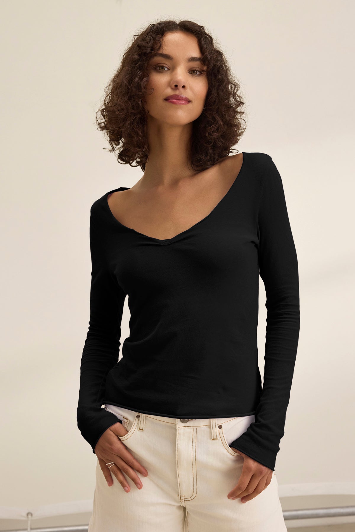 A woman with curly hair wears the NOURA TEE by Velvet by Graham & Spencer, a classic black long-sleeve V-neck in soft cotton jersey, paired with cream pants and standing against a plain light background.-39173154865345