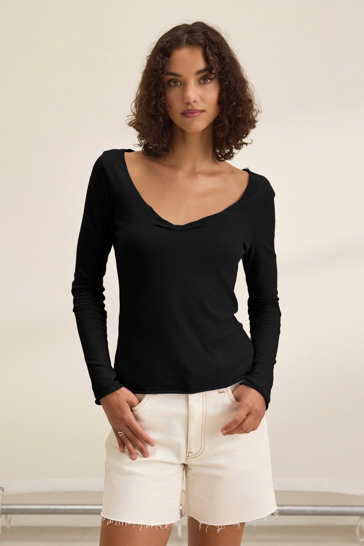 A woman with curly hair wears the NOURA TEE by Velvet by Graham & Spencer, a classic black long-sleeve cotton jersey top, paired with off-white high-waisted shorts, standing against a plain light background.-39173154898113