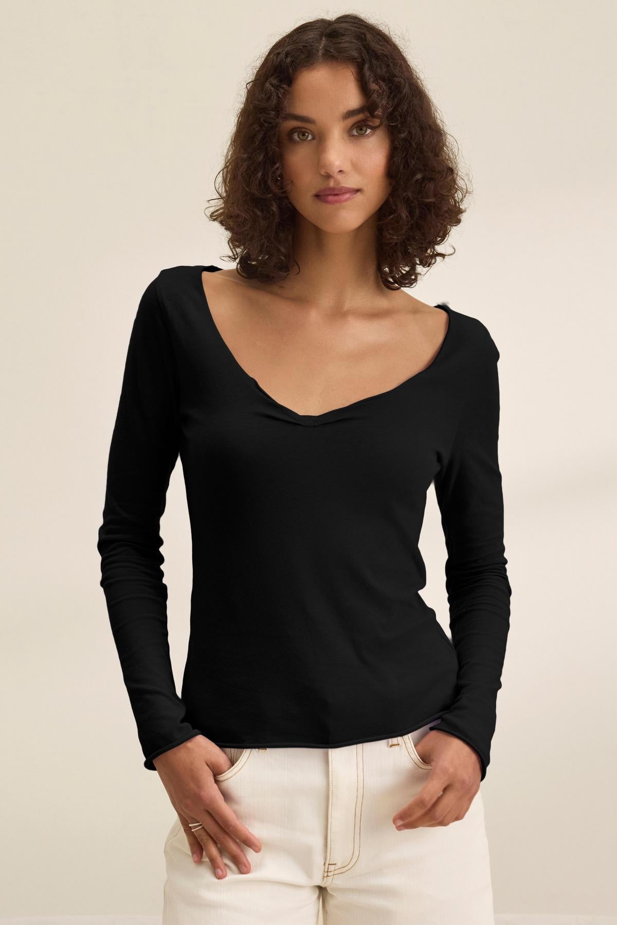 A woman with curly brown hair models the NOURA TEE by Velvet by Graham & Spencer—a black, long sleeve cotton jersey V-neck—paired with white pants against a plain light background.-40500868776129