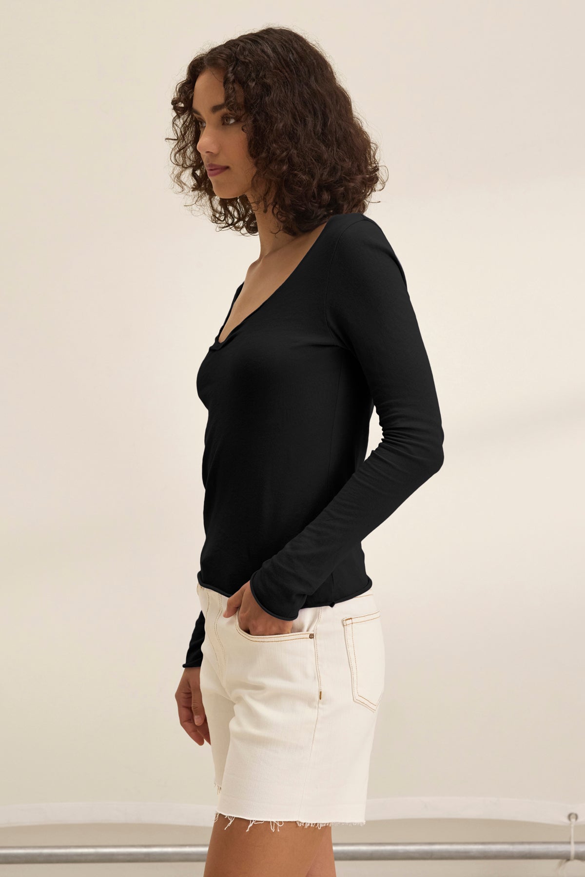   A woman with curly hair models the NOURA TEE by Velvet by Graham & Spencer, a classic fitted black long-sleeve cotton jersey top, paired with white denim shorts and standing sideways against a plain light background. 
