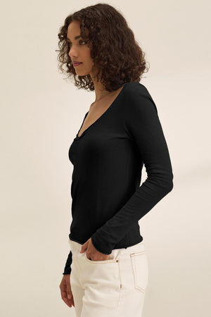 A woman with curly hair stands in side profile against a plain light background, wearing the NOURA TEE by Velvet by Graham & Spencer—a black long sleeve cotton jersey top—paired with white pants.