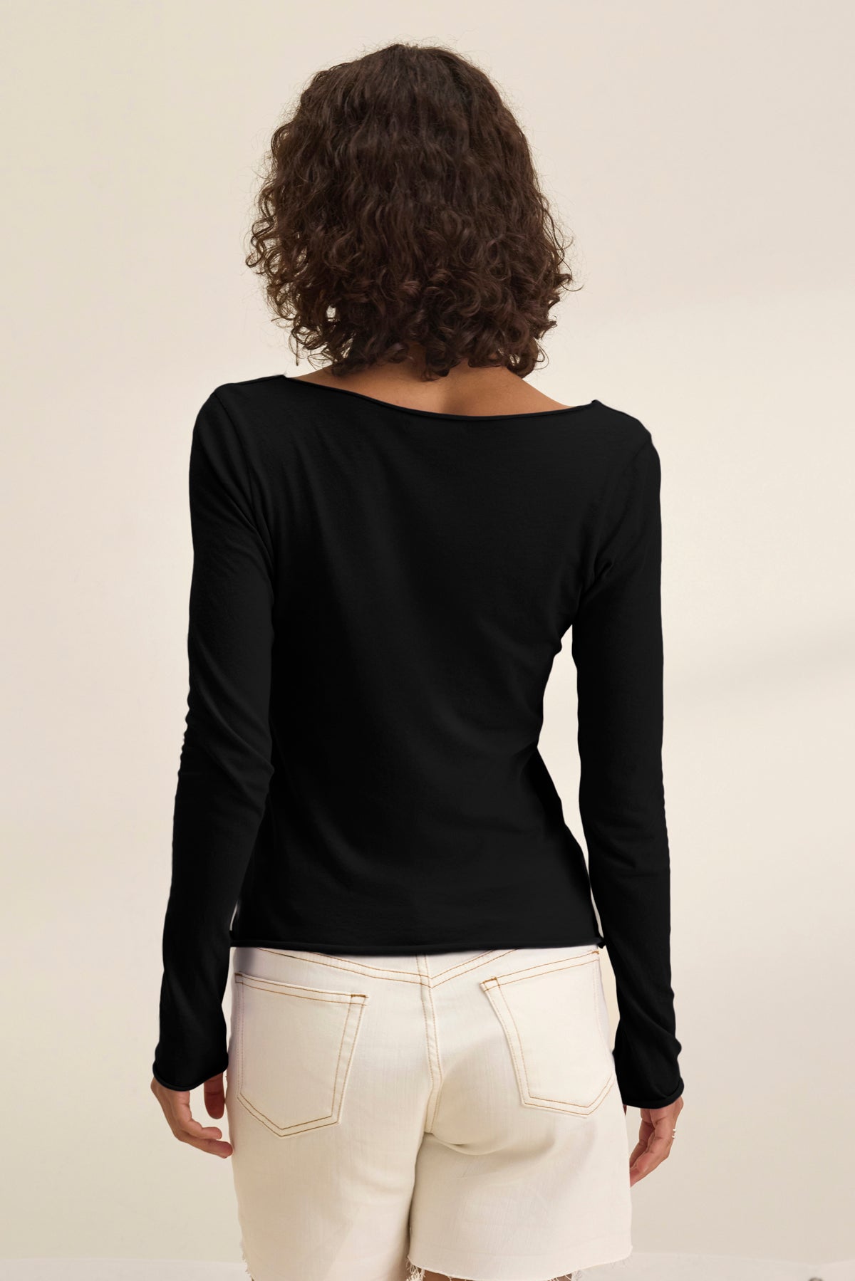 A person with curly brown hair is seen from the back wearing the Velvet by Graham & Spencer NOURA TEE, a classic black long-sleeve cotton jersey top, paired with white shorts against a plain light background.-39173154963649