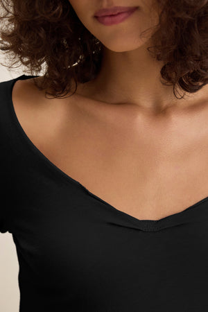 A woman with curly brown hair models the NOURA TEE by Velvet by Graham & Spencer, a classic black V-neck cotton jersey top, shown from the shoulders to just above the lips.