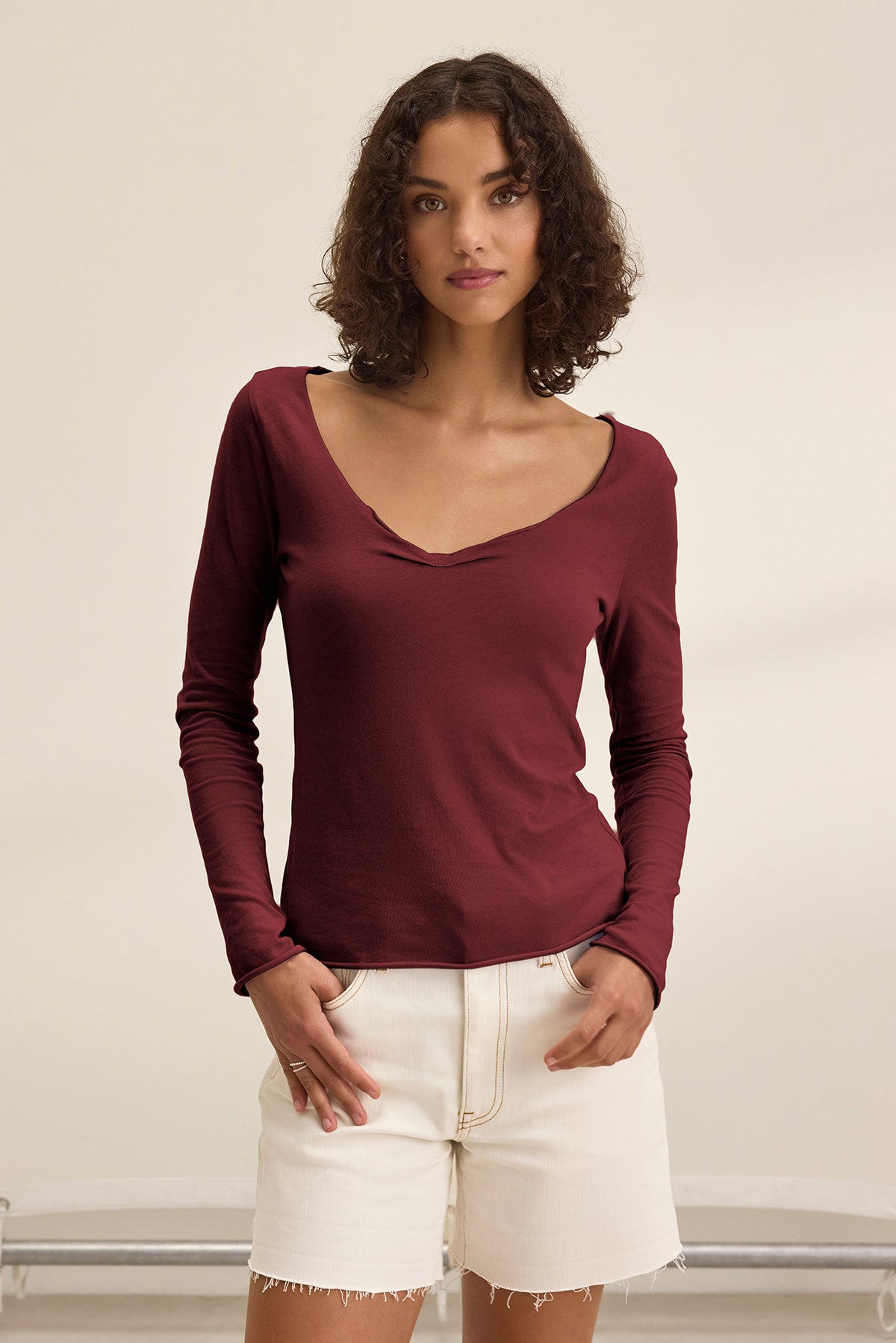 A woman with short curly hair wears the NOURA TEE by Velvet by Graham & Spencer—a long sleeve, deep burgundy cotton jersey with a scoop neckline—paired with off-white shorts, standing against a plain light background.-40009859498177