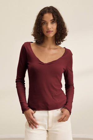 A woman with curly brown hair wears the NOURA TEE by Velvet by Graham & Spencer, a long-sleeve burgundy cotton jersey top, paired with off-white pants against a plain light background.