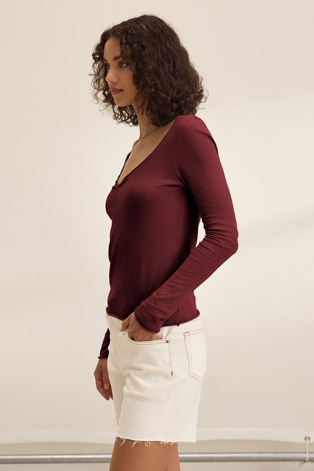A woman, shown in profile against a plain light background, wears the NOURA TEE by Velvet by Graham & Spencer— a long-sleeve scoop neck top styled with white denim shorts.-40009859596481