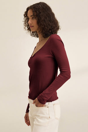 A person with curly brown hair wears the NOURA TEE by Velvet by Graham & Spencer— a long-sleeve cotton jersey top with a scoop neckline—paired with off-white pants, standing against a plain beige background.