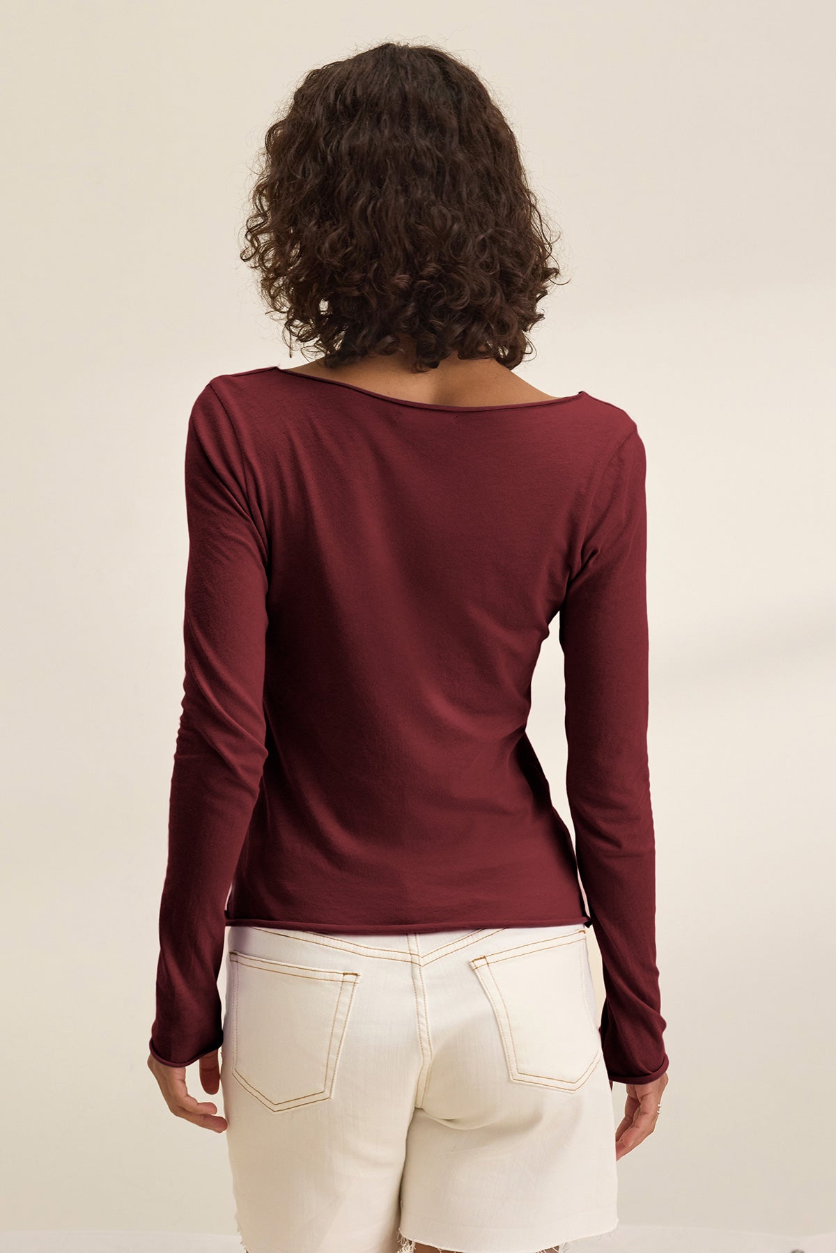 A person with curly hair, seen from behind, wears the NOURA TEE by Velvet by Graham & Spencer—a long-sleeve burgundy cotton jersey top with a scoop neckline—paired with high-waisted white shorts against a plain background.-40009859530945