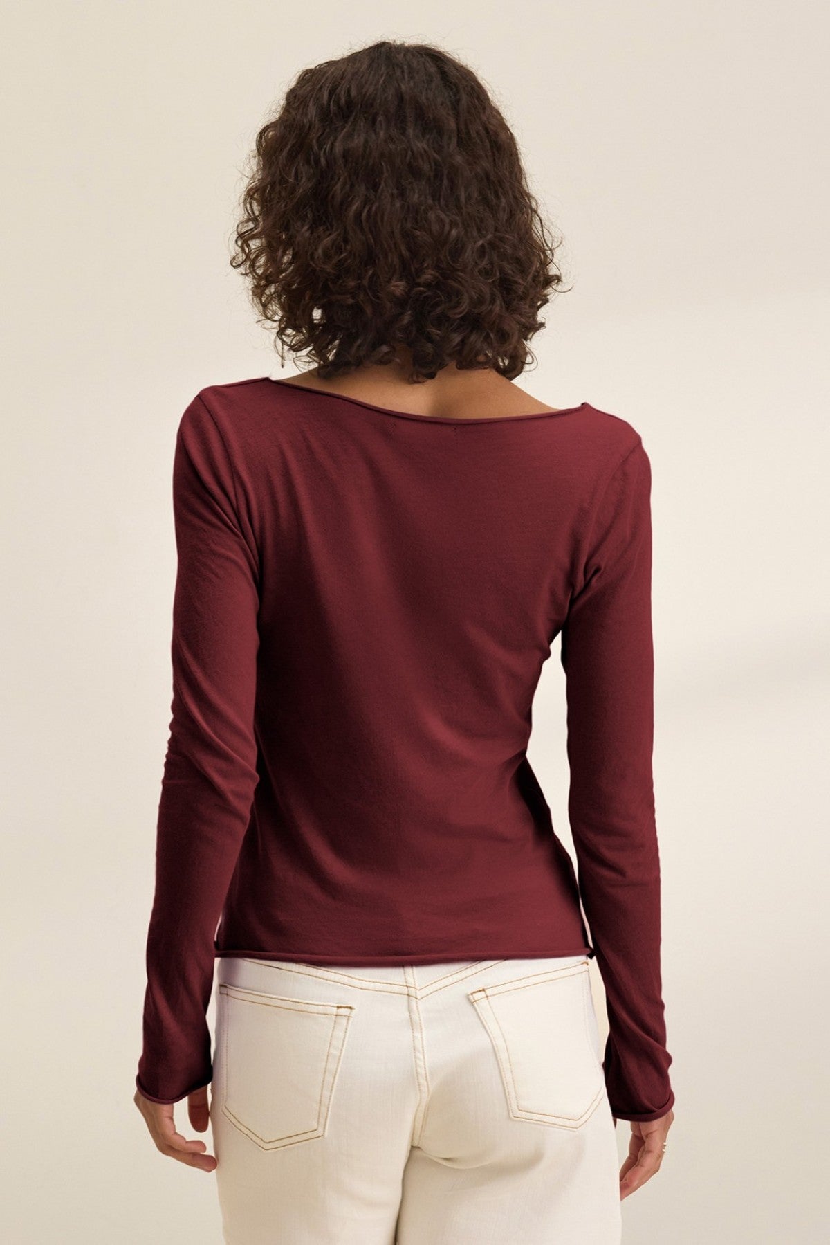A person with curly hair is shown from the back wearing Velvet by Graham & Spencer’s NOURA TEE, a long sleeve maroon cotton jersey top, paired with white pants against a neutral background.