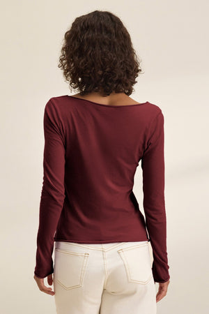 A person with curly hair is shown from the back wearing Velvet by Graham & Spencer’s NOURA TEE, a long sleeve maroon cotton jersey top, paired with white pants against a neutral background.