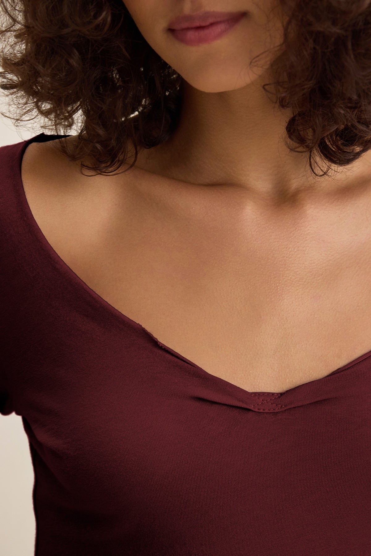 A woman with curly brown hair wears the NOURA TEE by Velvet by Graham & Spencer, a deep burgundy cotton jersey shirt with a scoop neckline, shown from her lips to her chest.-40009859563713