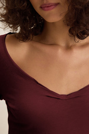 A woman with curly brown hair wears the NOURA TEE by Velvet by Graham & Spencer, a deep burgundy cotton jersey shirt with a scoop neckline, shown from her lips to her chest.