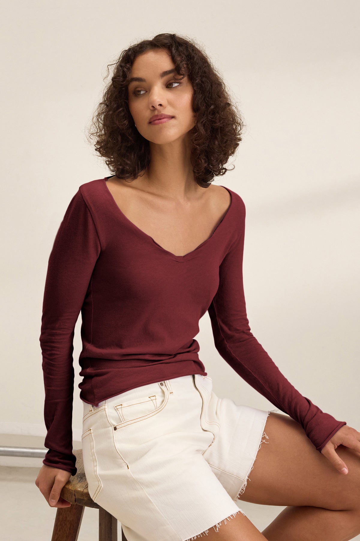 A woman with curly hair sits on a stool wearing the Velvet by Graham & Spencer NOURA TEE, a maroon long sleeve cotton jersey with a scoop neckline, paired with white frayed-edge shorts against a neutral background.-40009855303873