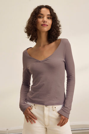 A person with curly brown hair models the Velvet by Graham & Spencer NOURA TEE, a timeless mauve, long-sleeved V-neck top, paired with cream pants against a plain light background.