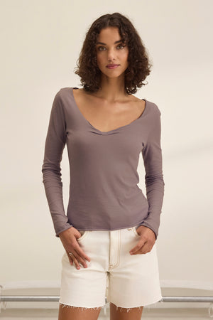 A woman with short curly hair wears the NOURA TEE by Velvet by Graham & Spencer, a classic long-sleeve taupe cotton jersey top, paired with white denim shorts. She stands against a plain light background with her hands in her pockets.