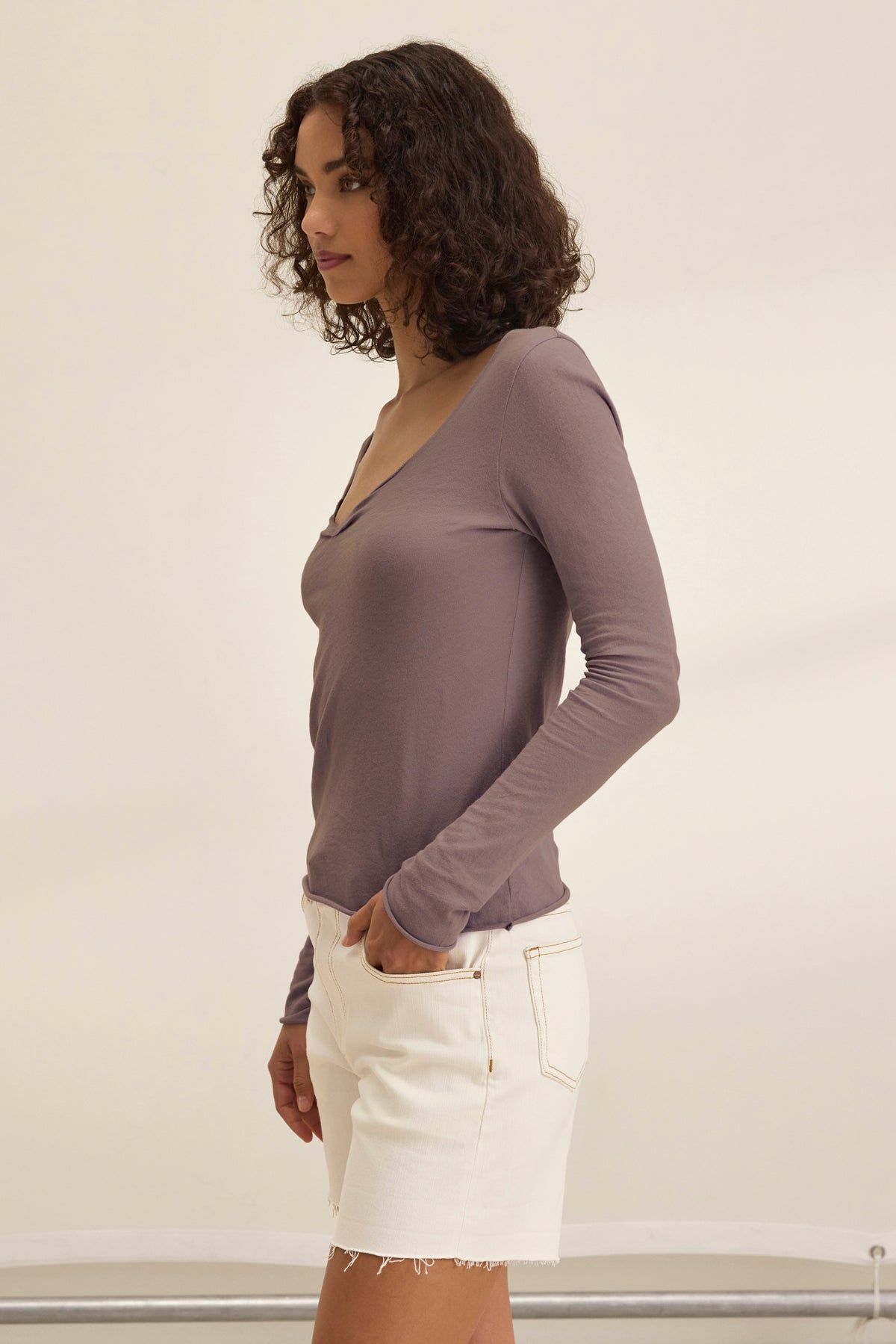 A woman with curly hair stands sideways, wearing the NOURA TEE by Velvet by Graham & Spencer—a classic mauve long-sleeve soft cotton jersey top—paired with white denim shorts against a plain, light background.-39173155094721