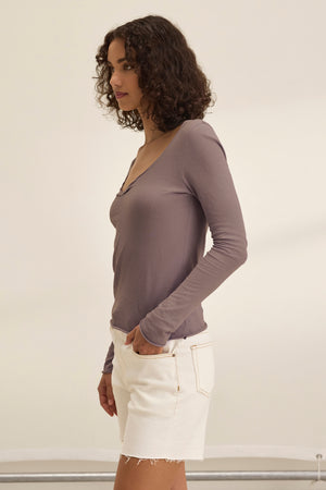 A woman with curly hair stands sideways, wearing the NOURA TEE by Velvet by Graham & Spencer—a classic mauve long-sleeve soft cotton jersey top—paired with white denim shorts against a plain, light background.