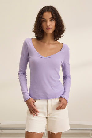 A person with curly hair, showcasing a classic style in the NOURA TEE by Velvet by Graham & Spencer and white shorts, stands against a plain background.