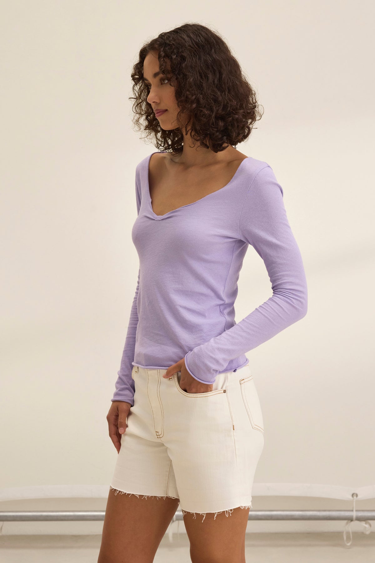 A woman with curly hair, exuding timeless charm, wears a Velvet by Graham & Spencer NOURA TEE in light purple and white shorts. She stands indoors, gazing thoughtfully to the side.-38928710369473