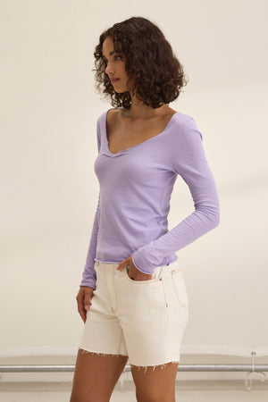 A woman with curly hair, exuding timeless charm, wears a Velvet by Graham & Spencer NOURA TEE in light purple and white shorts. She stands indoors, gazing thoughtfully to the side.
