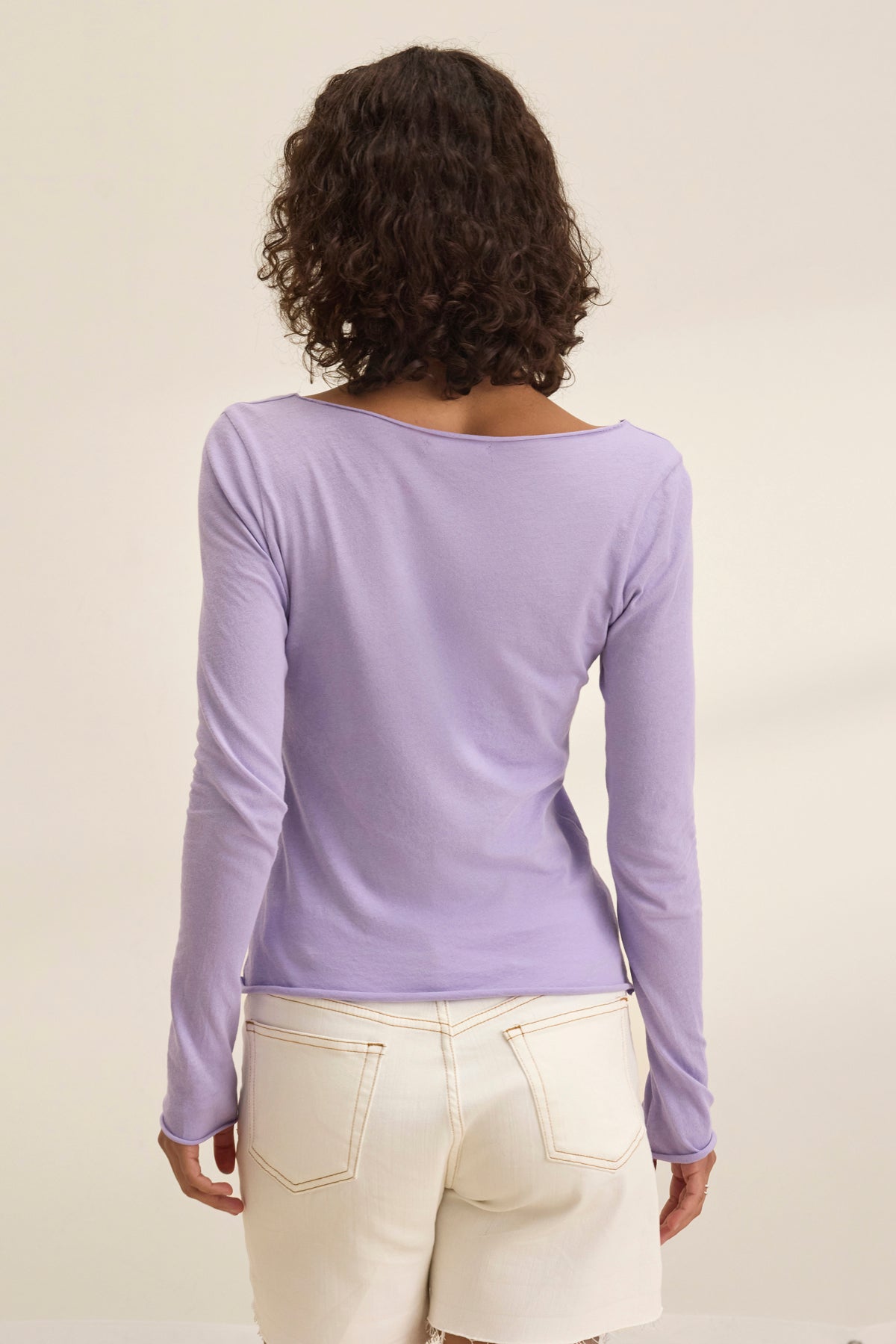 A person with curly hair, shown from behind, wears the timeless NOURA TEE by Velvet by Graham & Spencer—a long-sleeved lavender shirt—paired with white pants against a plain background.-38928710336705