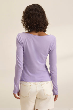 A person with curly hair, shown from behind, wears the timeless NOURA TEE by Velvet by Graham & Spencer—a long-sleeved lavender shirt—paired with white pants against a plain background.
