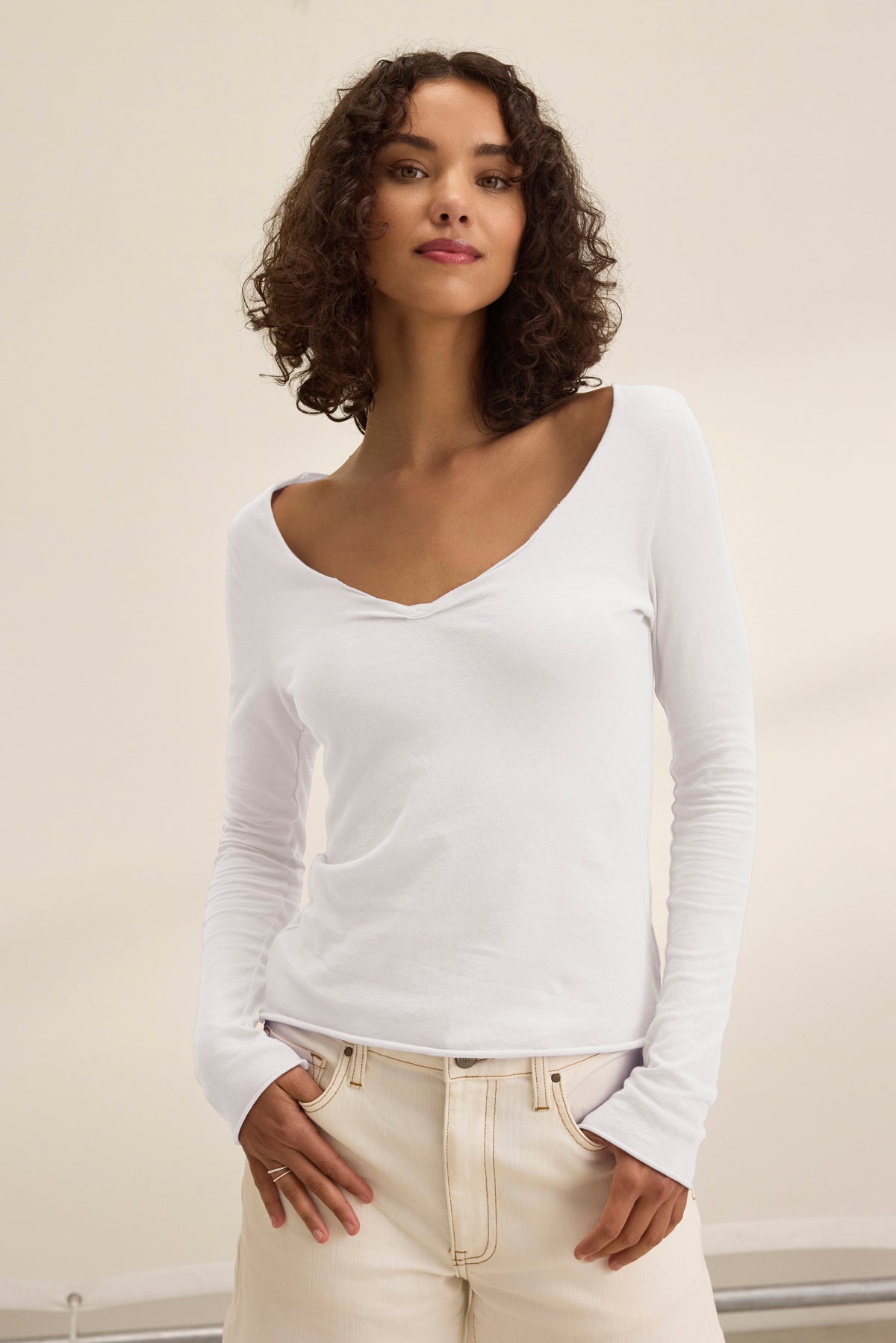 A person with curly hair stands against a plain background wearing the NOURA TEE from Velvet by Graham & Spencer, a classic white V-neck long-sleeve shirt made of soft cotton jersey, paired with light-colored pants.-38954020995265