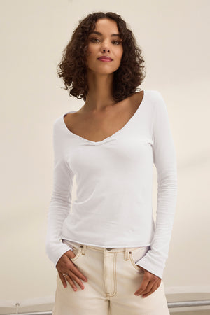 A person with curly hair stands against a plain background wearing the NOURA TEE from Velvet by Graham & Spencer, a classic white V-neck long-sleeve shirt made of soft cotton jersey, paired with light-colored pants.
