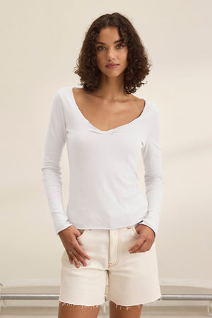 Dressed in a classic ensemble, a model showcases Velvet by Graham & Spencer's NOURA TEE, a soft cotton jersey long-sleeve top, paired with beige shorts against a plain backdrop.