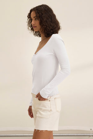 A person with curly hair is wearing the NOURA TEE, a white cotton jersey long-sleeve top from Velvet by Graham & Spencer, and beige shorts, standing sideways against a plain background.