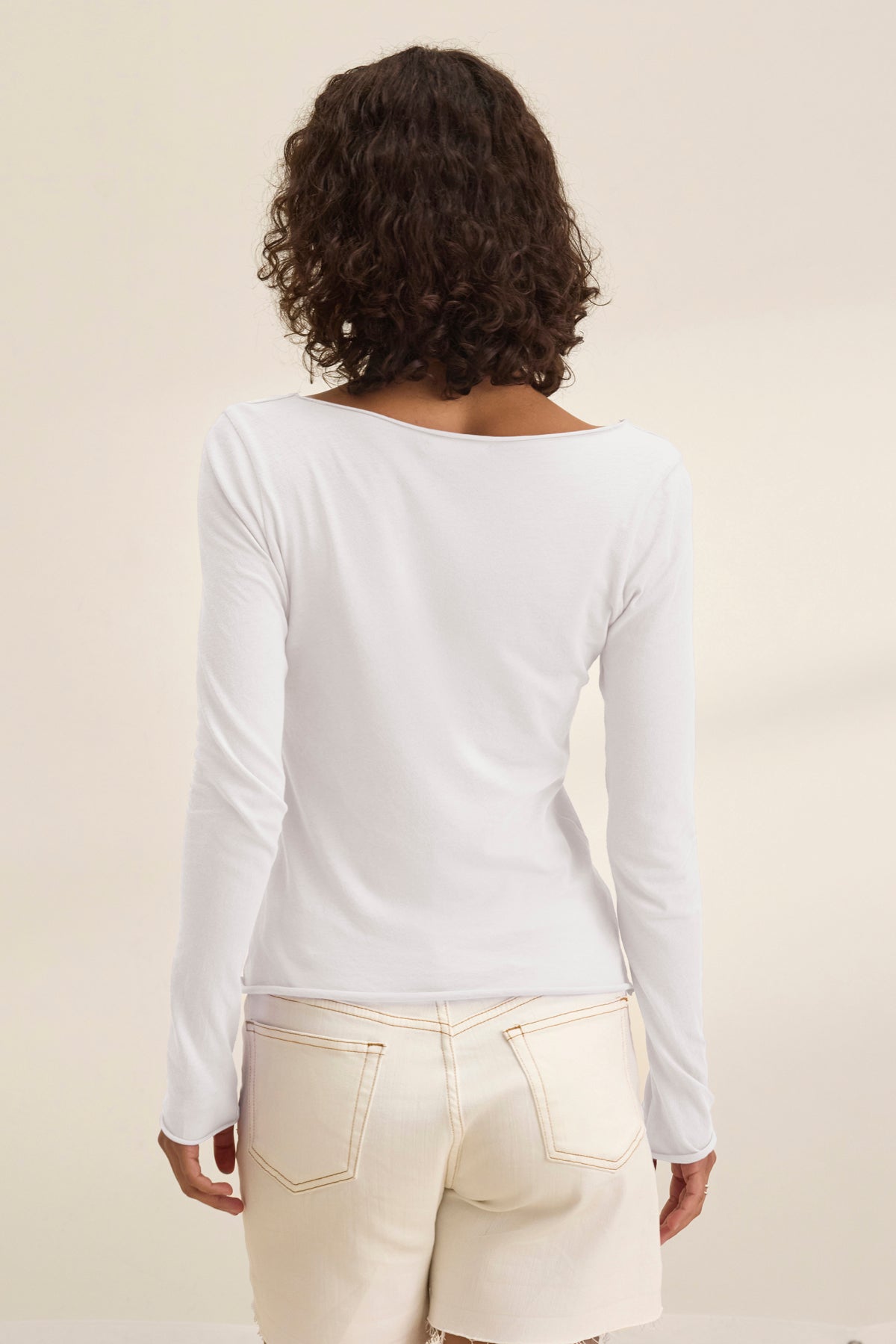 Person with curly hair wearing Velvet by Graham & Spencer's NOURA TEE in a long-sleeved white cotton jersey paired with beige pants, seen from behind.-38954021126337