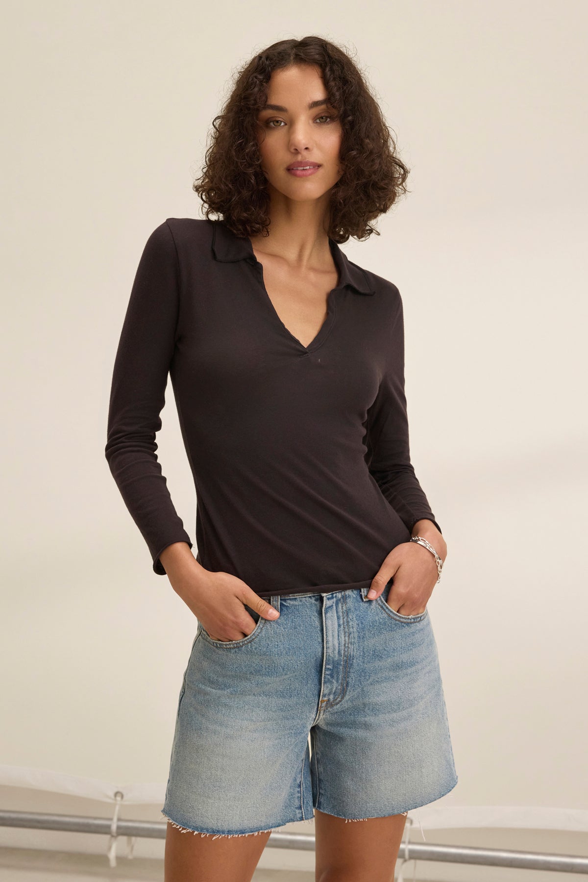 A woman with curly hair, wearing the Velvet by Graham & Spencer REED POLO in black and denim shorts, stands confidently with hands in pockets against a neutral background, showcasing a classic style that echoes timeless collections.-38928814145729